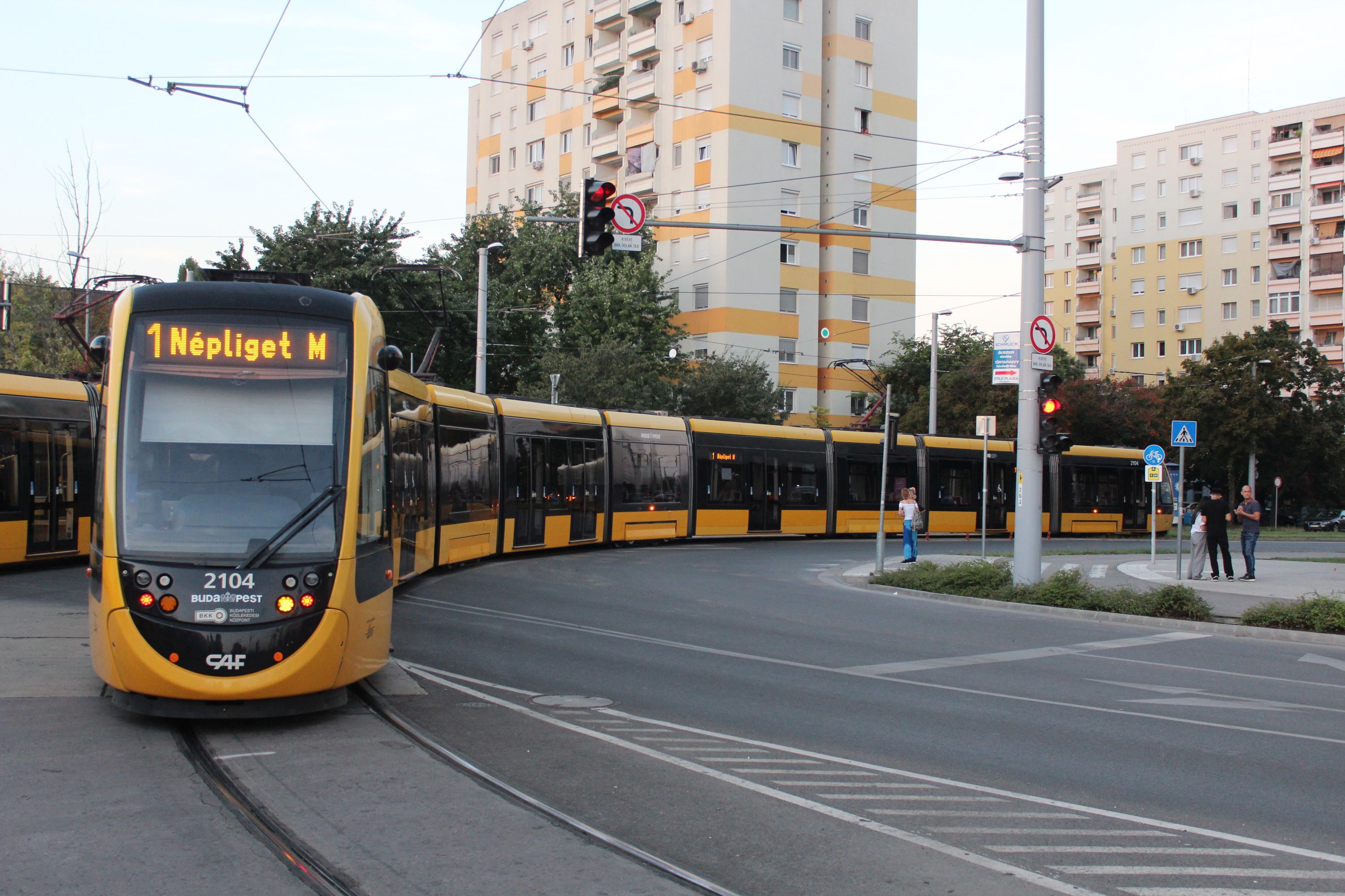 CAF Urbos 3 (9-członowy) #2104 na linii 1 skręca w Etele út po odjeździe z przystanku początkowego Kelenföld vasútállomás w Budapeszcie (25 sierpnia 2024)